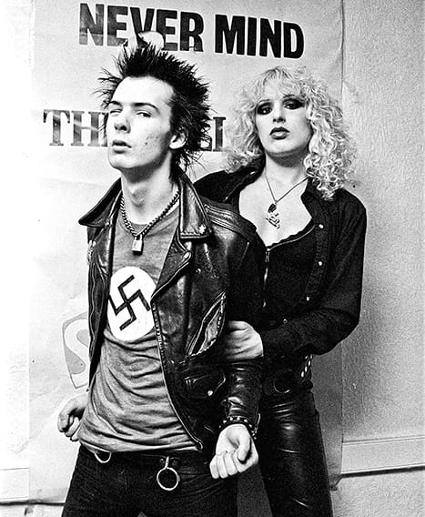 Sid Vicious and Nancy Spungen. Vicious is scowling and wearing a leather jacket and under it a swastika t-shirt, while Spungen stands behind him, holding his arms as if to hold him back. On the wall behind them is a poster for Never Mind the Bollocks, Here's the Sex Pistols.